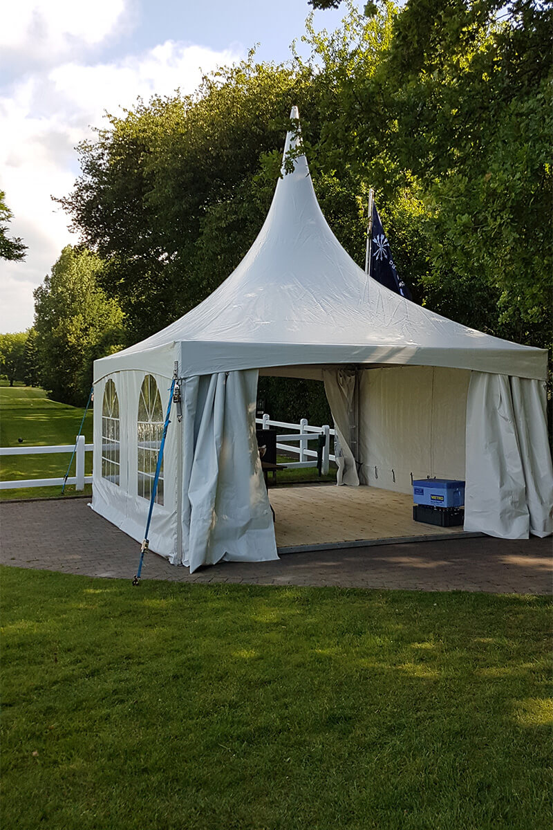 Tente blanche en forme de pagode installée sur une pelouse, avec un drapeau et un environnement verdoyant.